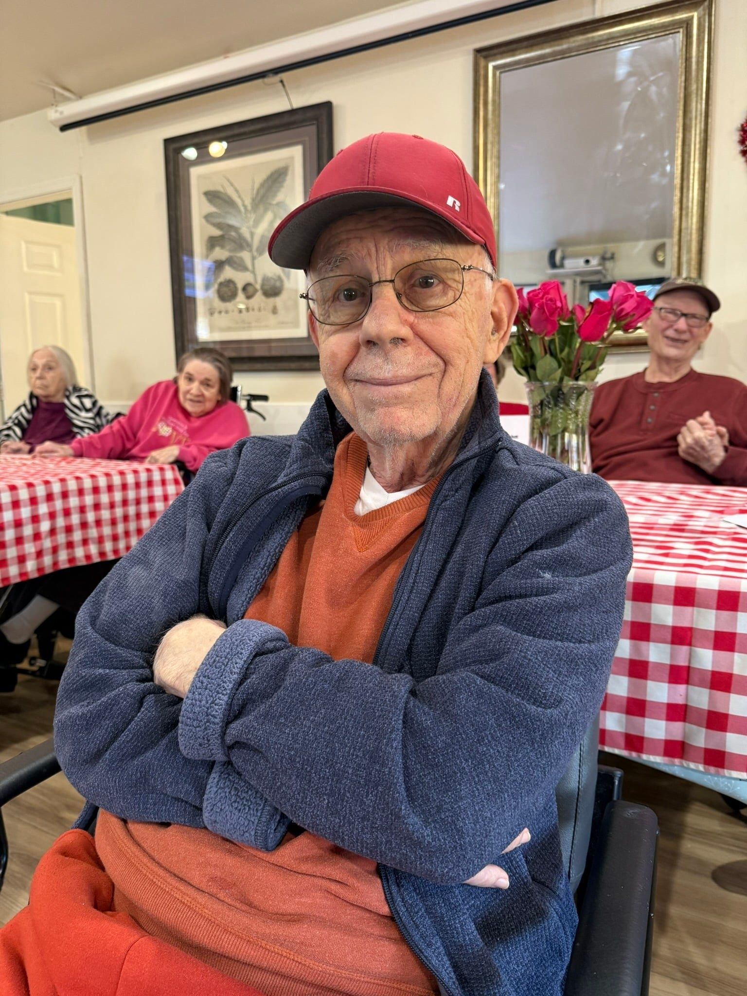 Man in red cap and orange sweater sits with arms crossed at a table in a community dining room.