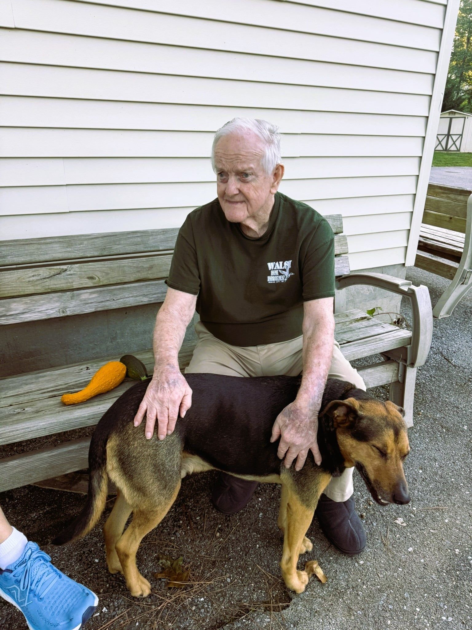 Man sitting on a bench petting a large brown-and-black dog beside him on a porch.