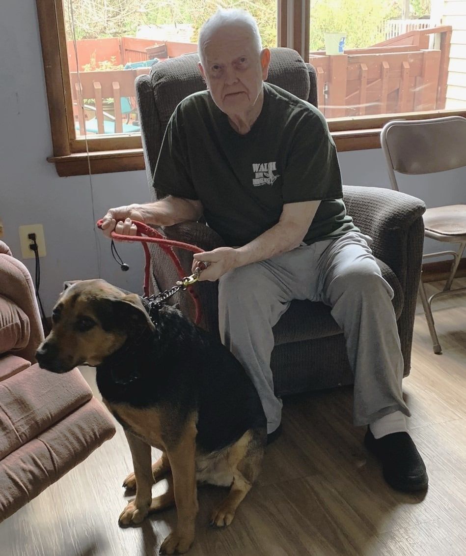 Man sitting in a chair indoors, holding a red leash attached to a black-and-tan dog in the foreground
