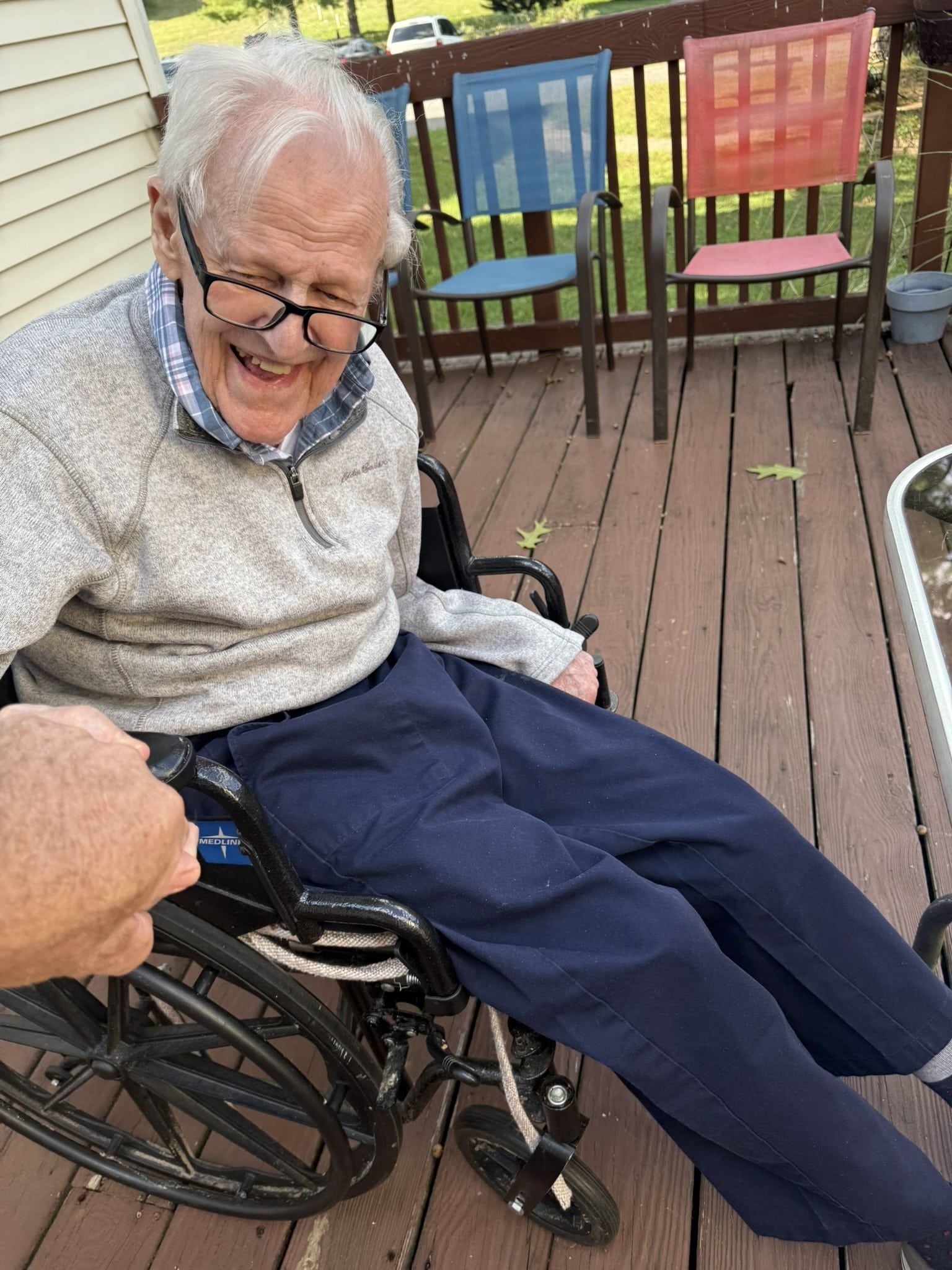 Elderly person smiling in a wheelchair on a wooden deck, with colorful chairs behind them.