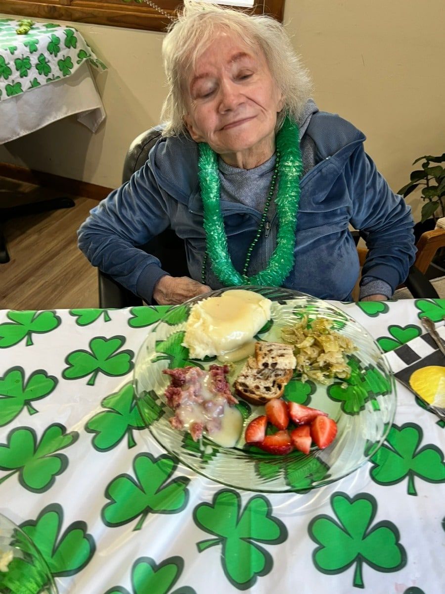 Woman at a table with a shamrock tablecloth, smiling beside a plate of food and green beads.