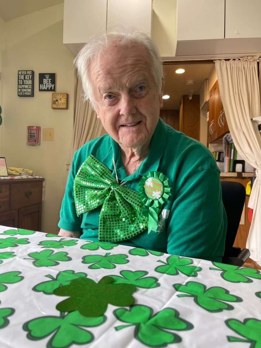 Elderly person in a green outfit with a shamrock bow sits at a table decorated with shamrocks.