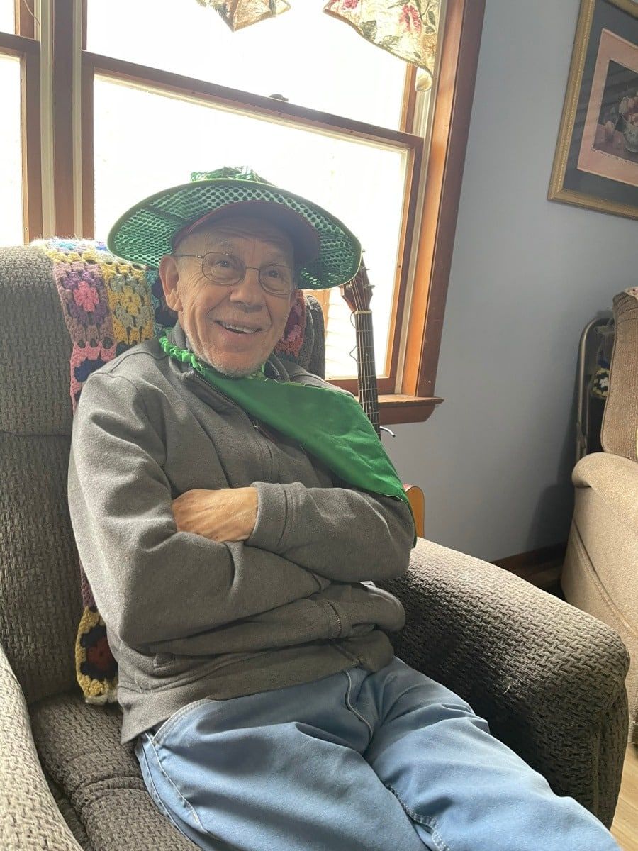 Smiling person seated in an armchair, wearing a green hat and scarf beside a bright window
