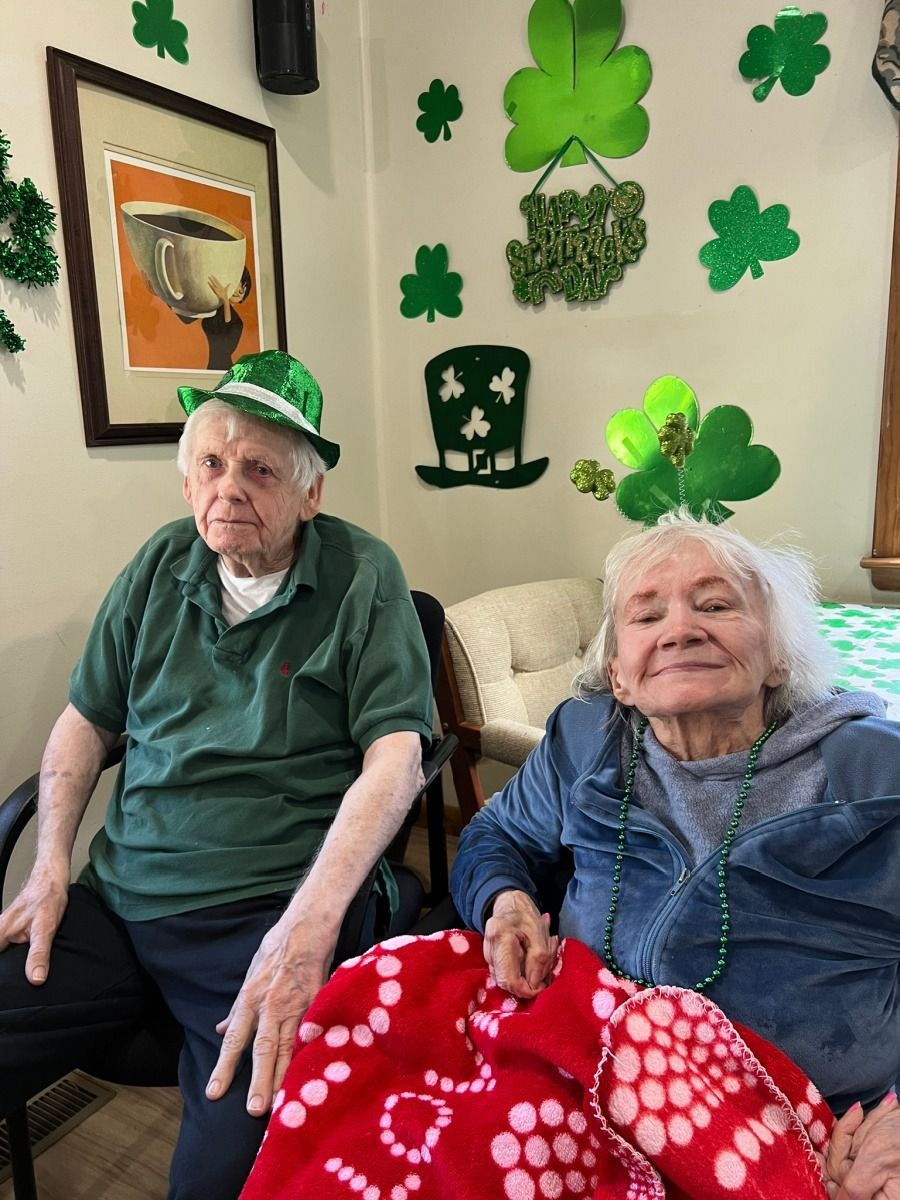 Two elderly people sit indoors with St. Patrick’s Day decorations on the wall and hats on their heads.