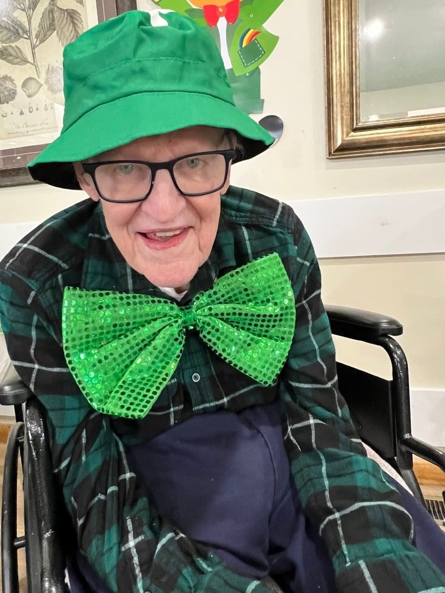 Smiling person in a green hat and bow tie, seated in a wheelchair indoors.