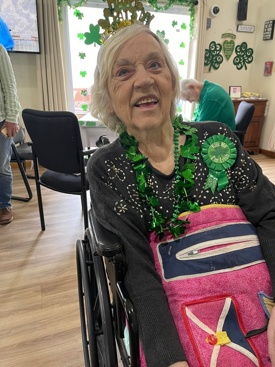 Elderly woman smiling in a wheelchair, wearing a green lei and colorful blanket in a decorated room.