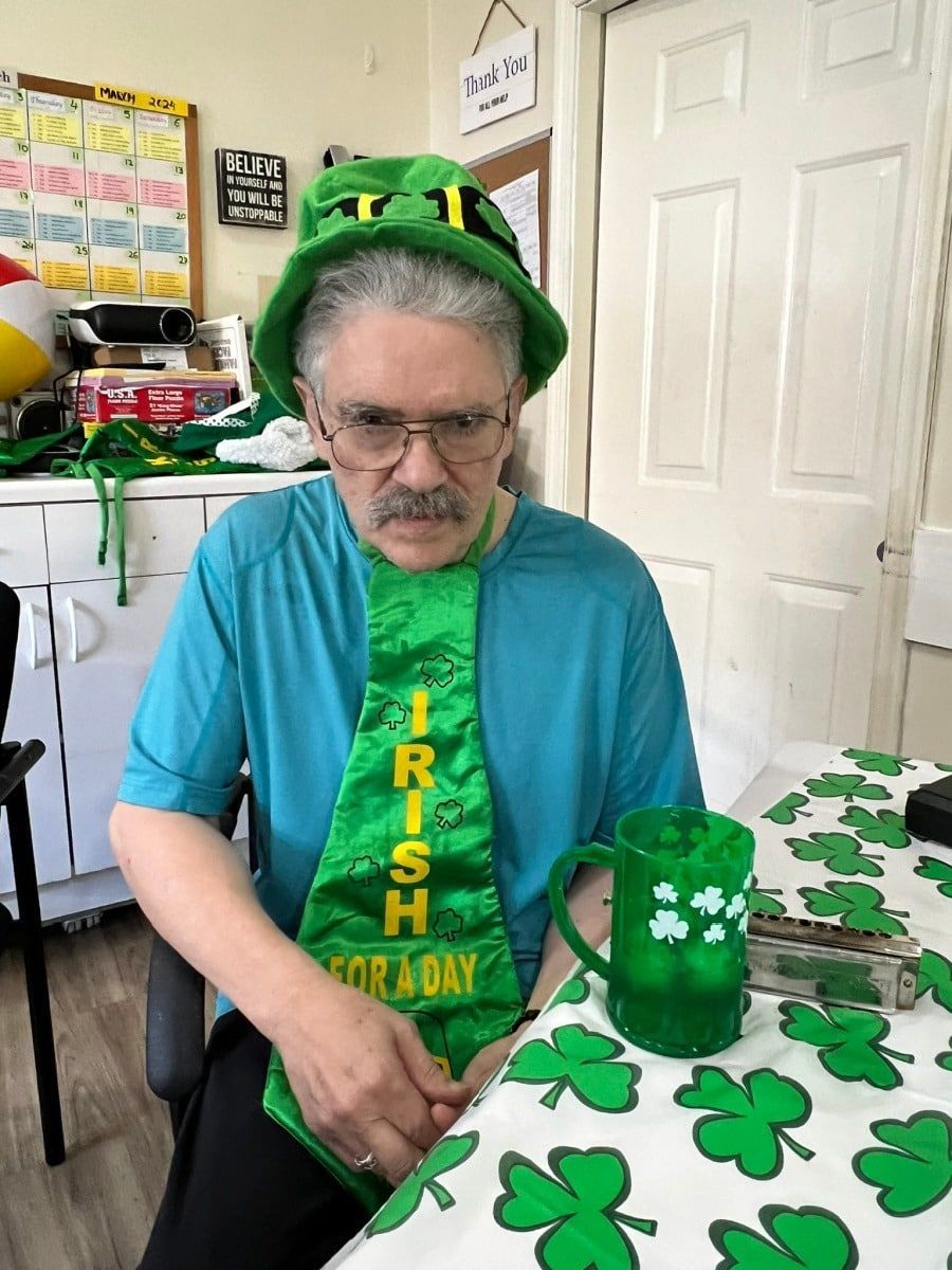 Person in a green St. Patrick’s Day outfit and hat, sitting at a table with shamrock decorations and a green cup.