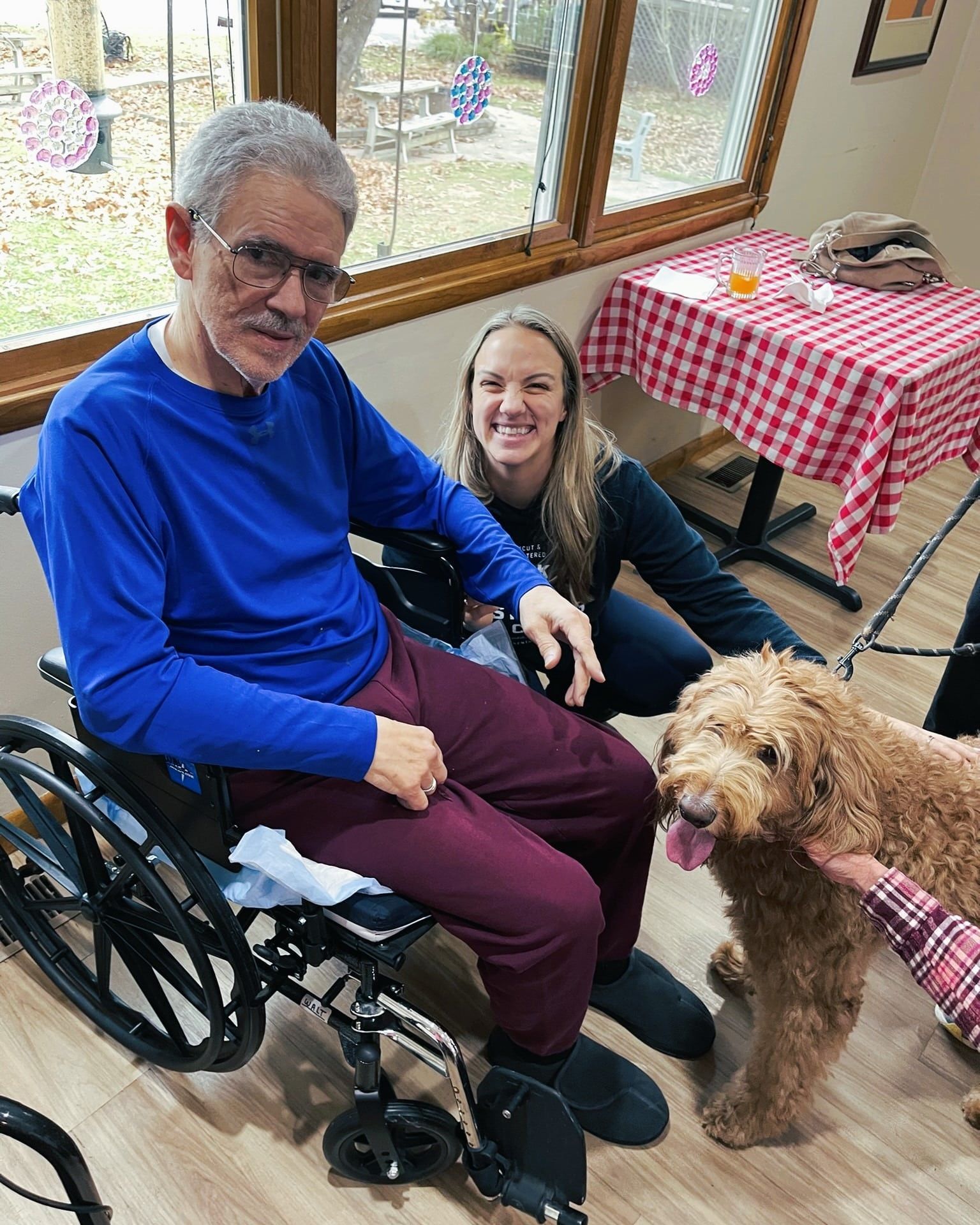Man in wheelchair petting a fluffy dog with a smiling woman beside him in a bright room with checkered tablecloths