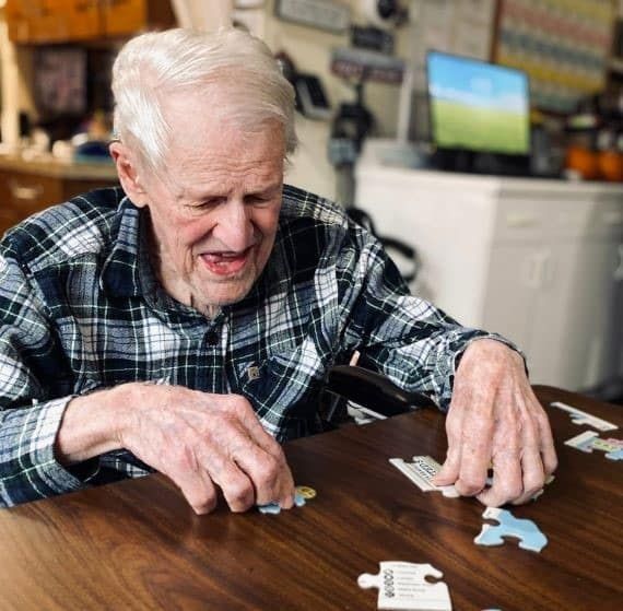 Elderly man in a plaid shirt playing dominoes at a table, smiling in a cozy room