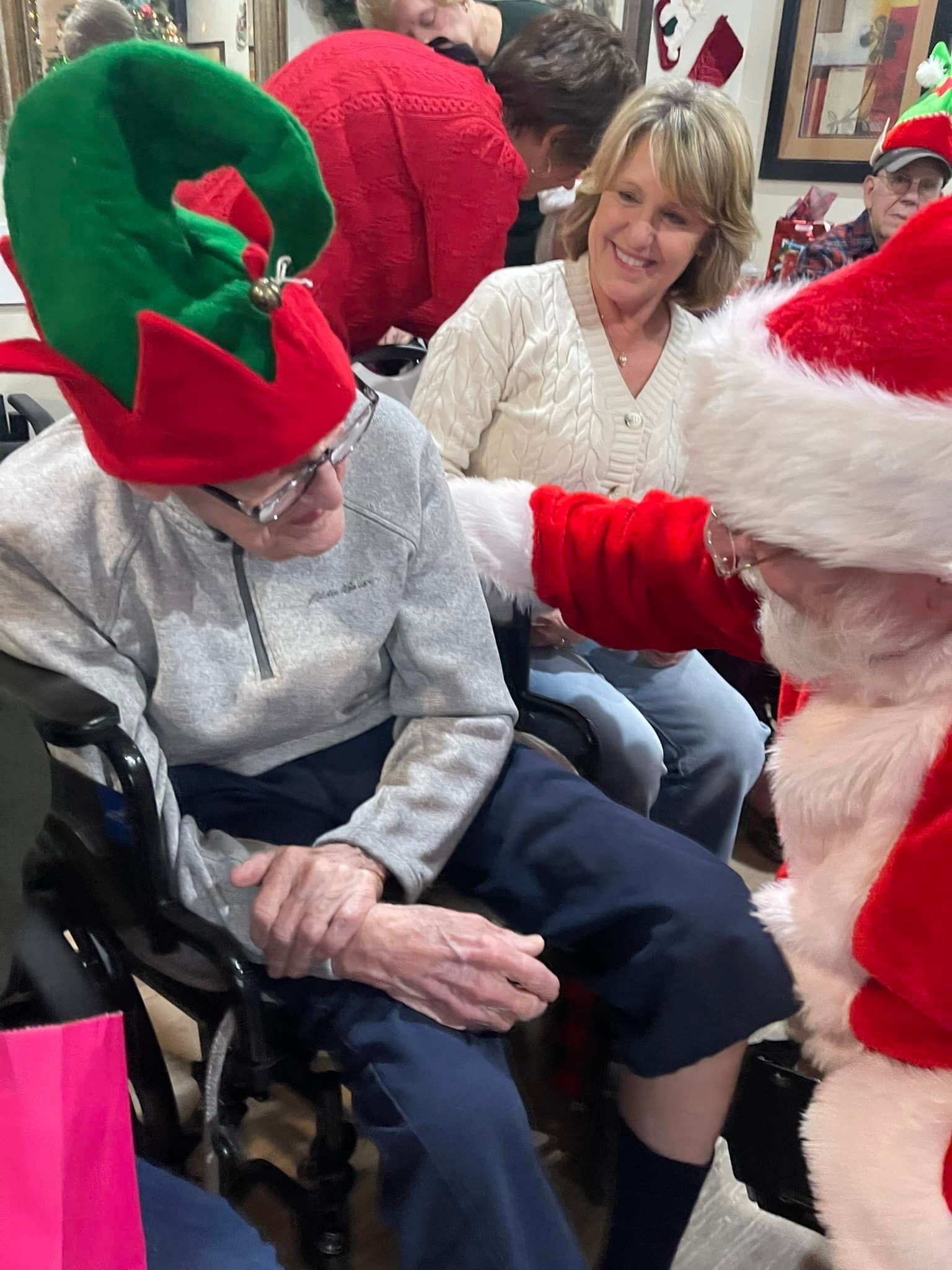 Woman in festive hat smiling with a seated person as Santa reaches out in a holiday setting