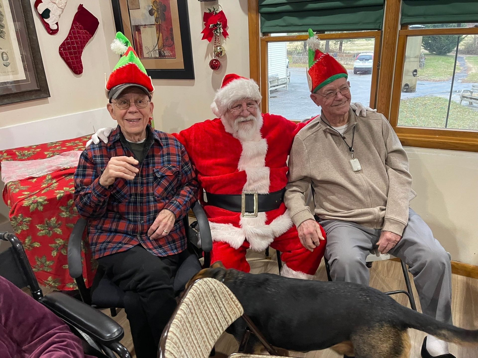 Three people in festive elf hats sit with Santa by a decorated fireplace, with a black dog in front.