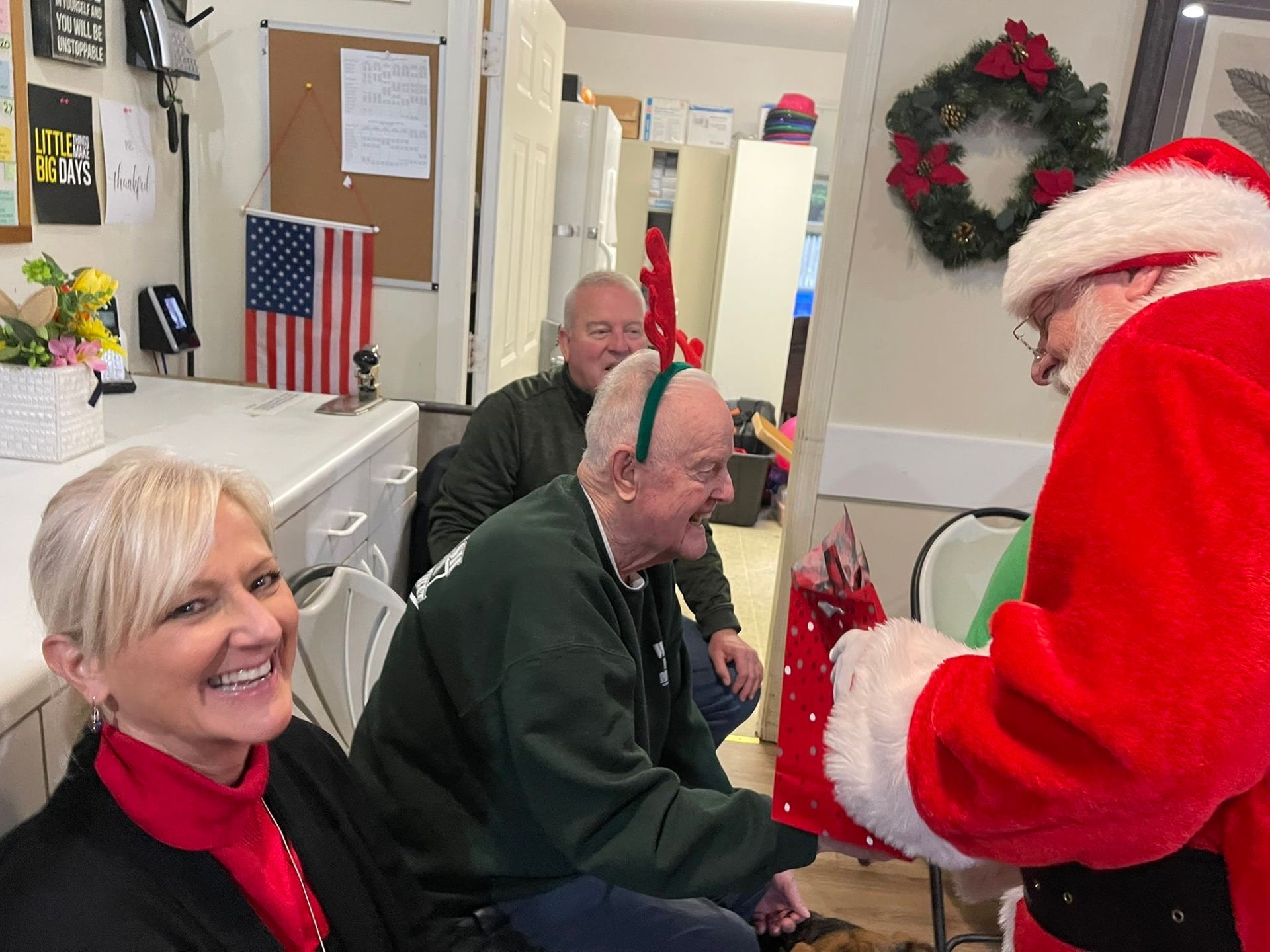 Santa handing a toy to a smiling person in a decorated room with Christmas wreaths and lights