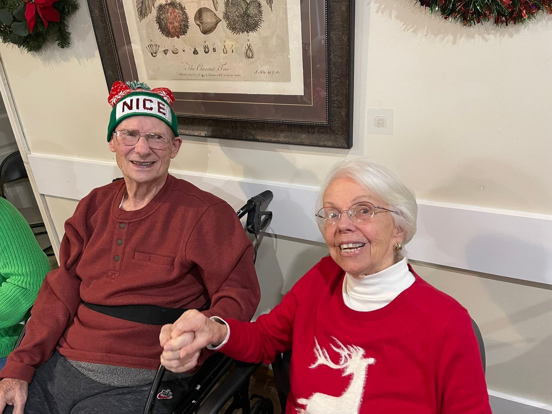 Two smiling people in festive sweaters seated indoors, one wearing a “NICE” holiday hat.