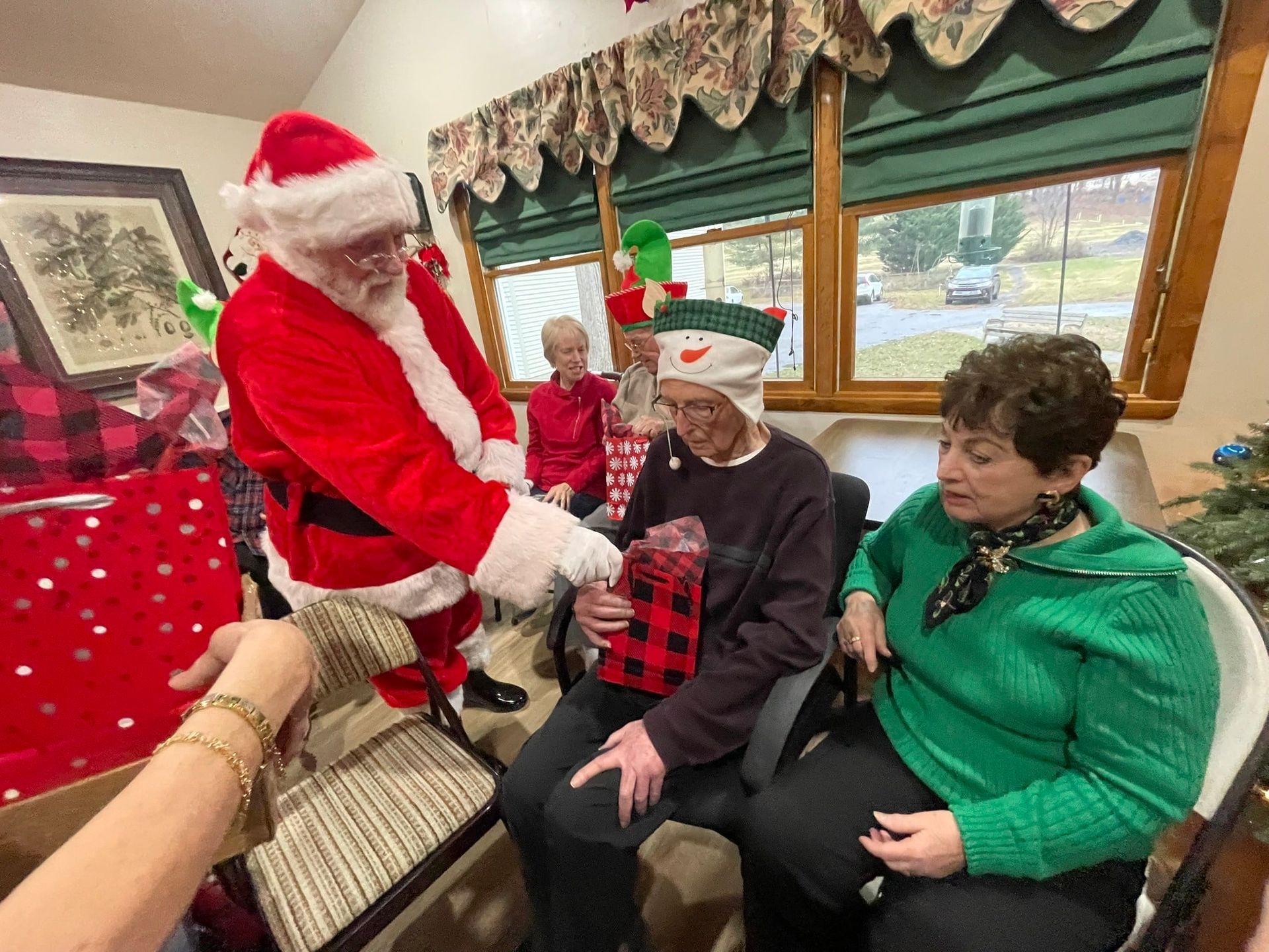 Santa hands a red gift to a seated person in a festive room with holiday decorations.