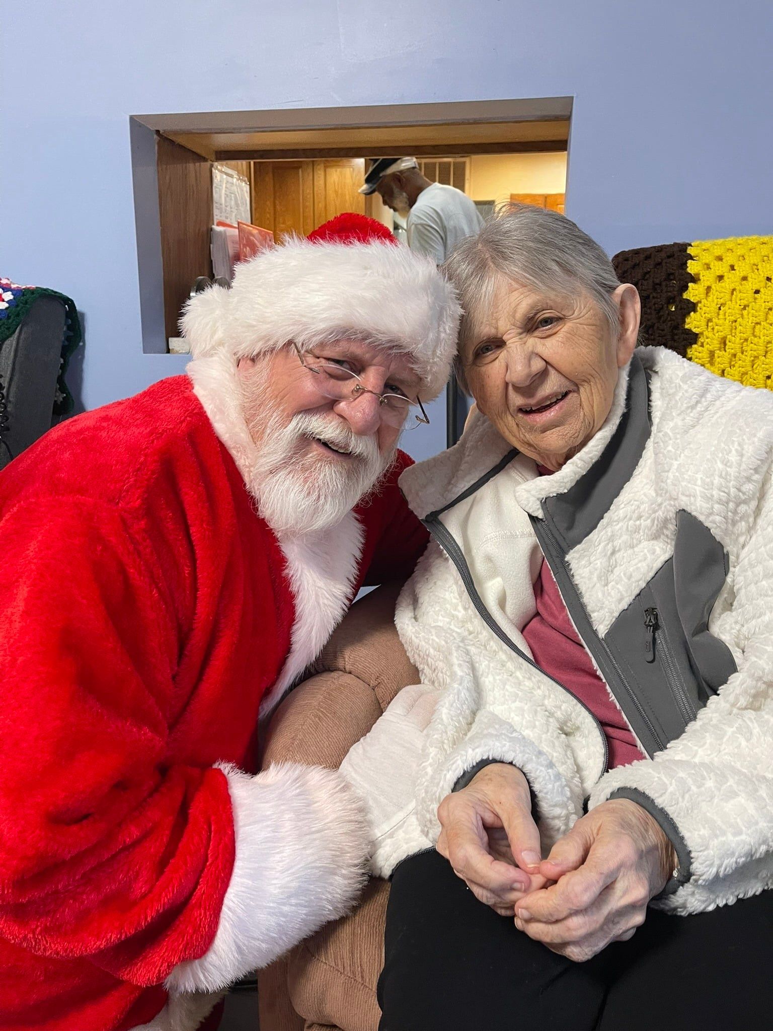 Santa sits beside an elderly woman indoors, smiling and leaning close for a festive photo.
