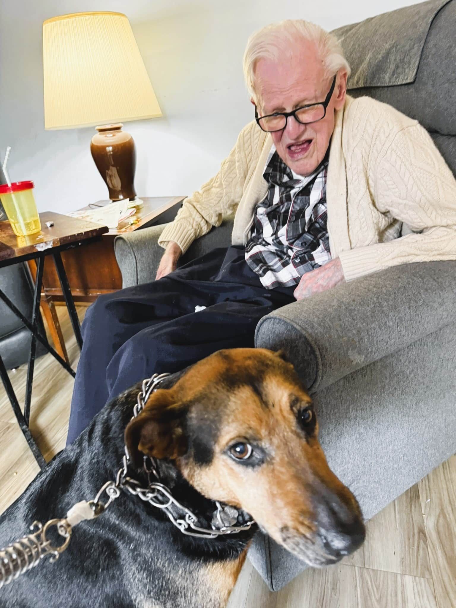 Older man seated on a couch with a large dog resting in front, in a cozy living room with a lamp nearby
