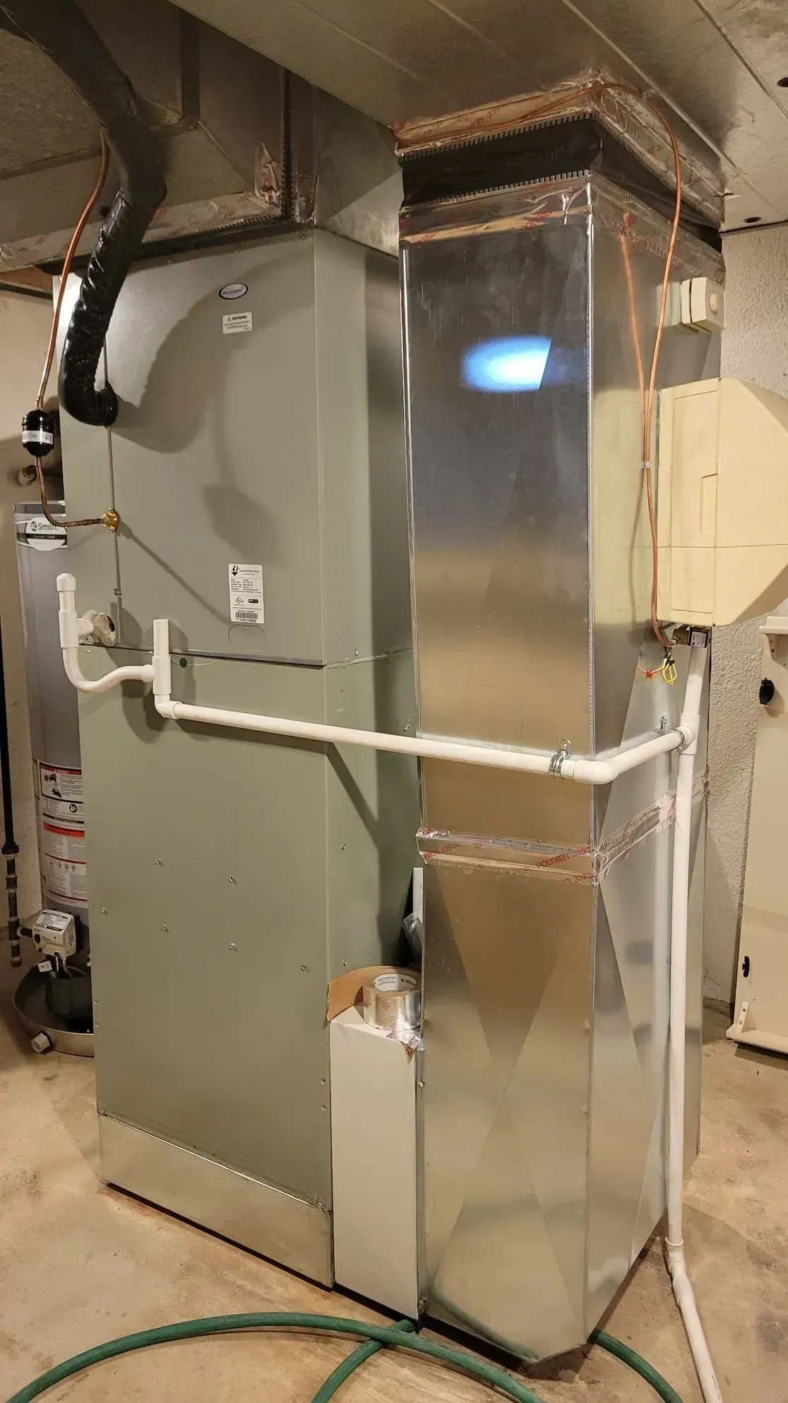 A stainless steel air conditioner is sitting in a basement next to a green hose.
