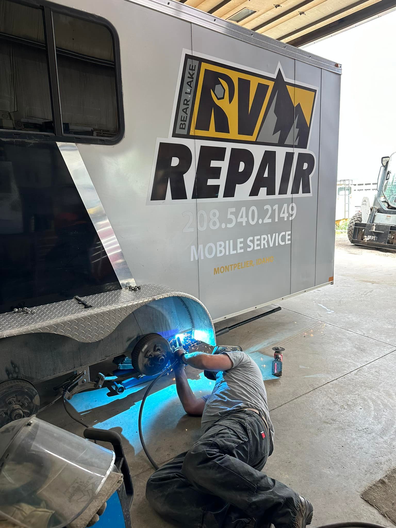 Man welding under an RV; RV repair shop with logo visible in background.