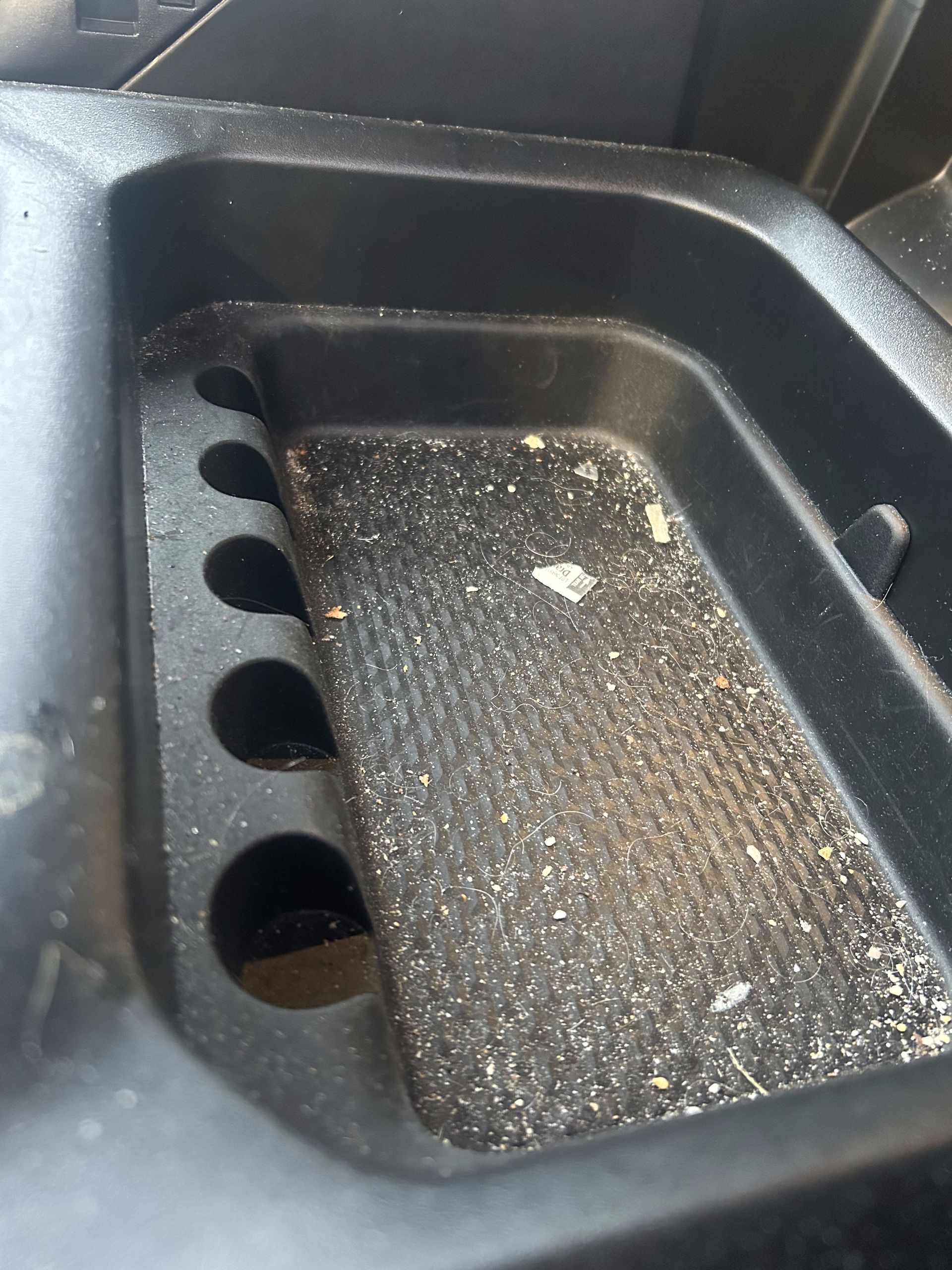 Black, dusty tray in a vehicle interior with several circular holes and textured bottom.