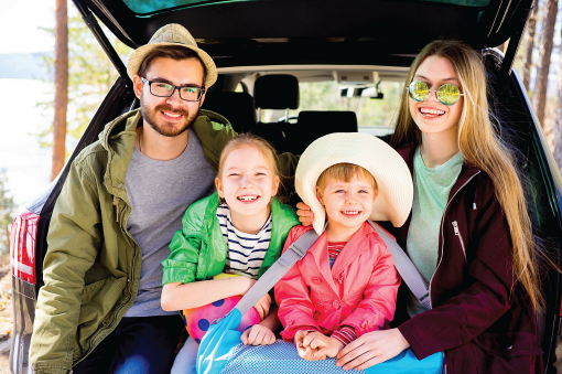 Family of four smiles inside a car's open trunk in a forest setting; they are ready for a trip.