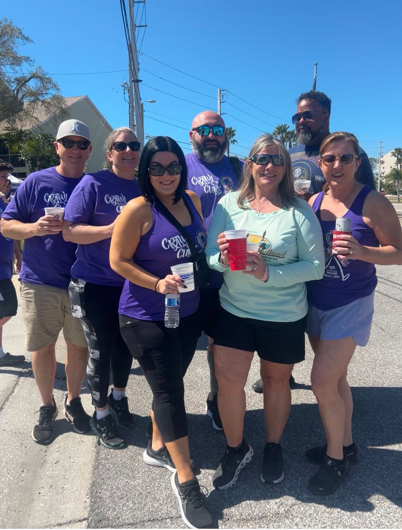Group of people in purple shirts, holding drinks, smiling outdoors in bright sunlight.