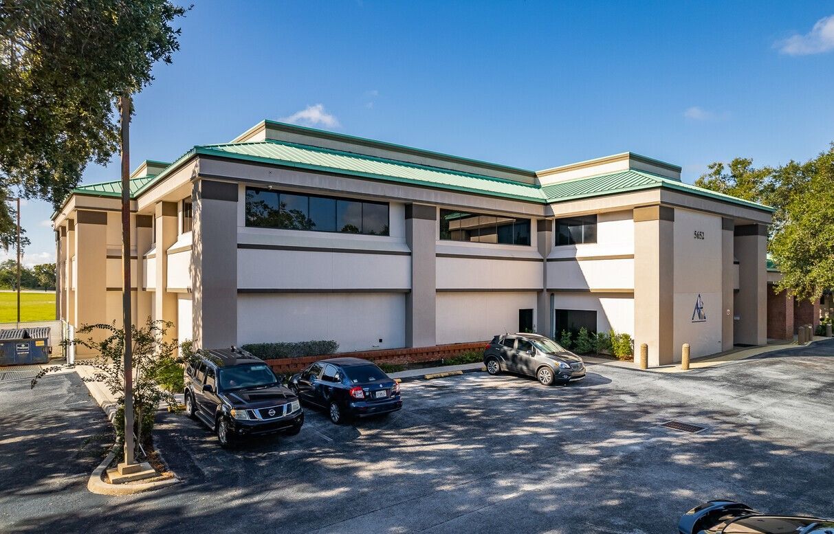 Office building with parked cars on a sunny day; green roof, beige walls, and blue sky.