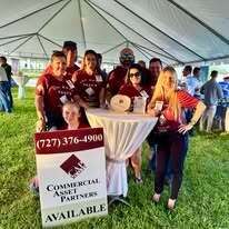 Group of people under tent, Commercial Asset Partners sign, smiling faces, green lawn.