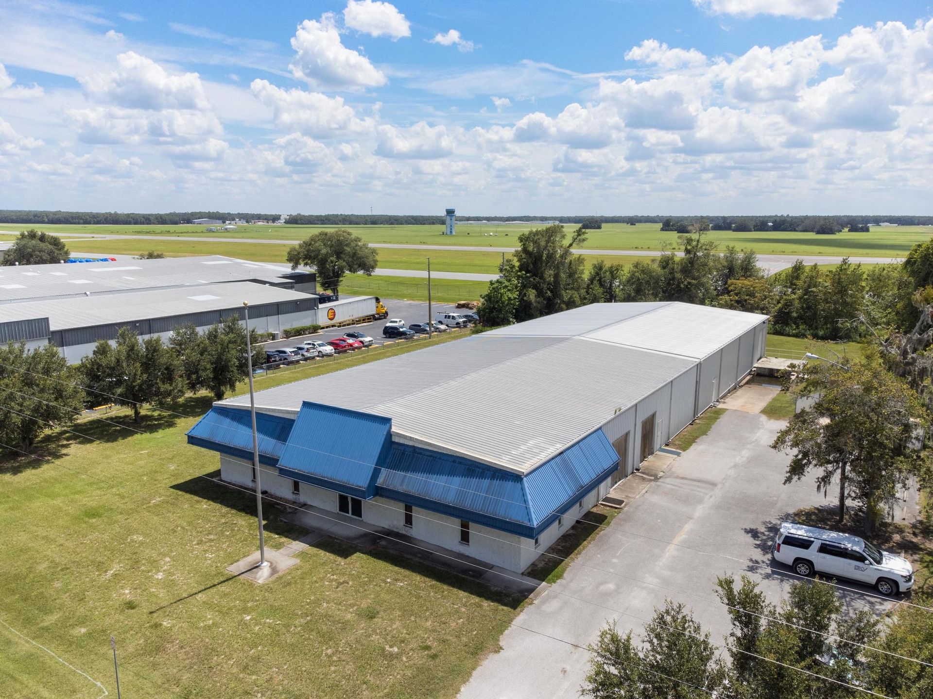 Aerial view of a warehouse with blue accents on the roof. A few vehicles and trees are visible.