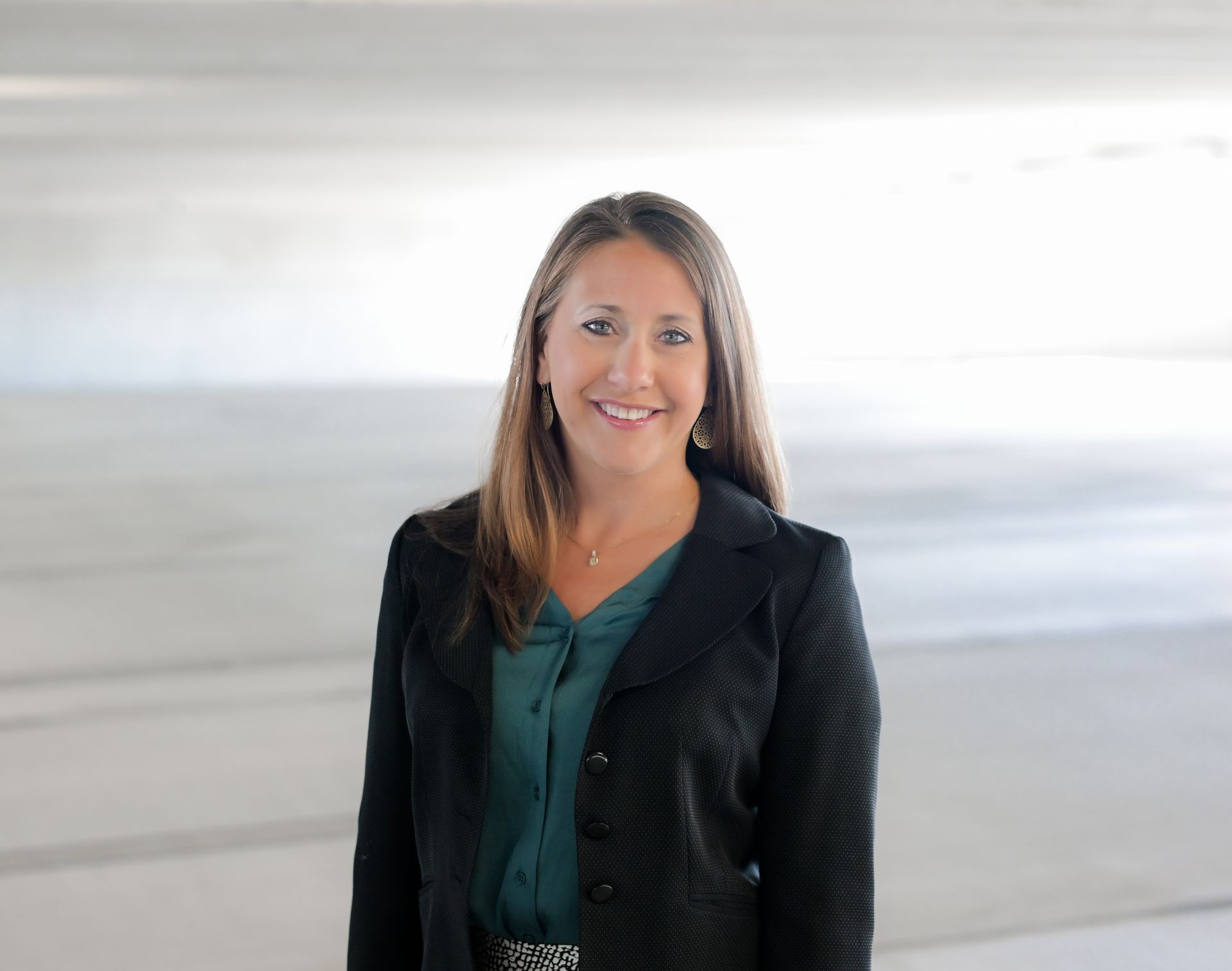 Woman with brown hair in a dark blazer and teal blouse smiles, posing in a bright, concrete space.