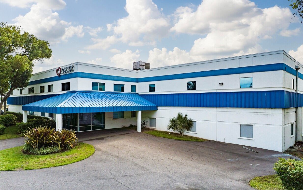 Blue and white building with entrance canopy, logo on roof. Driveway and greenery in foreground.