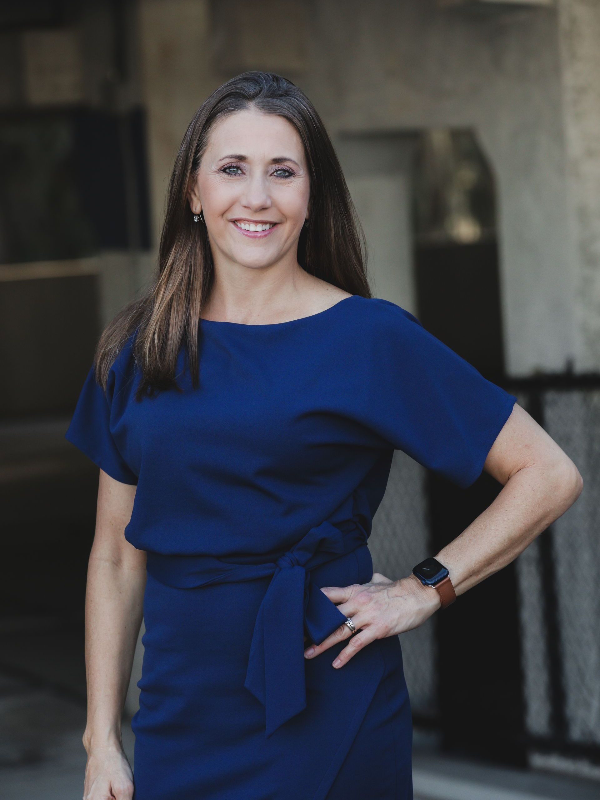 Woman with dark hair, wearing a blue dress, smiles and poses with her hand on hip.