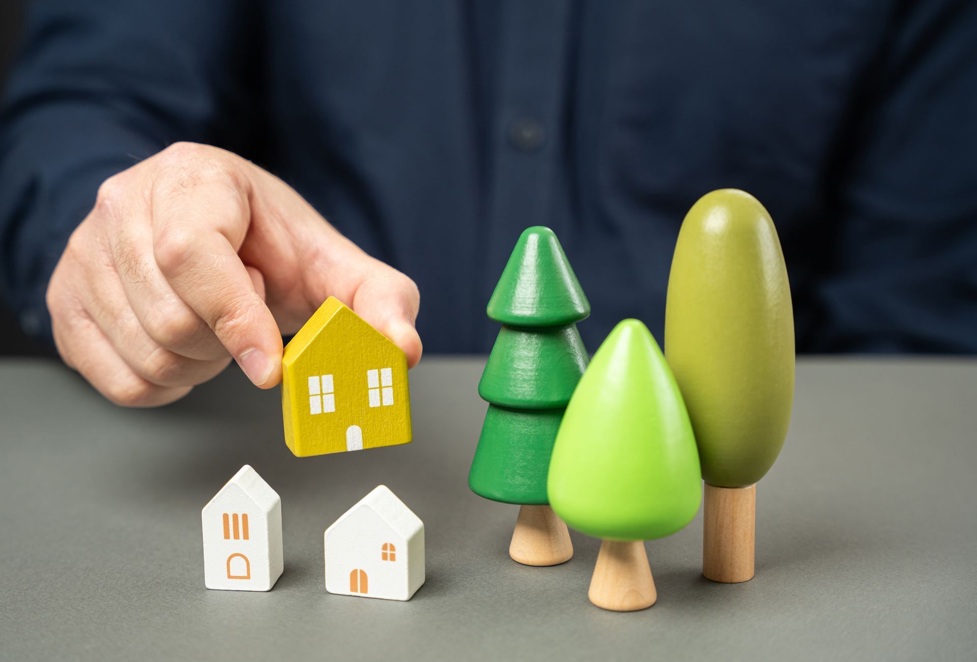 Hand holding a yellow toy house beside small white houses and green toy trees on a gray surface