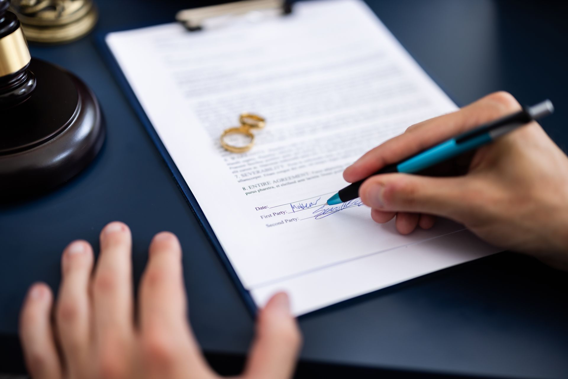 Hand signing a document on a desk beside a wedding ring and stamp, close-up.