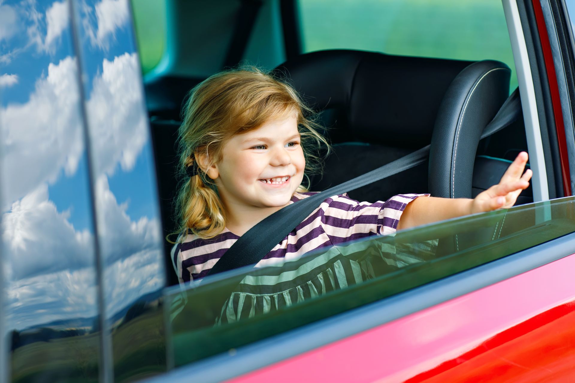 Young child in a car seat waving from a red car, smiling, with a blue sky reflecting in the window.