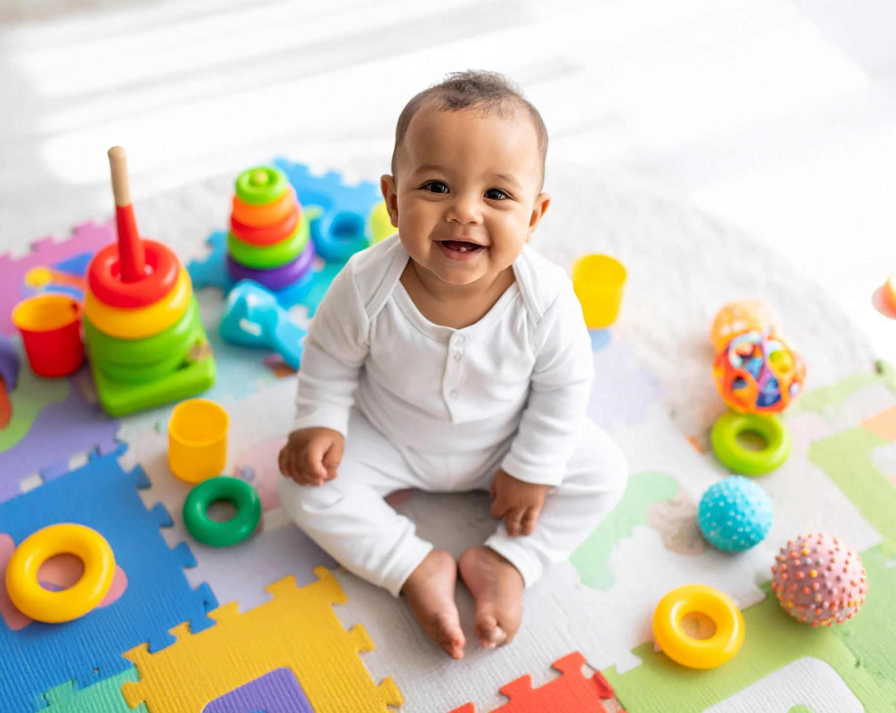 Smiling baby surrounded by colorful toys on a play mat.