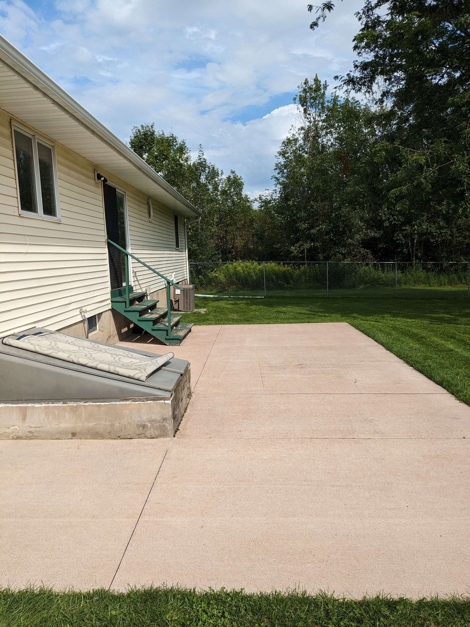A light-colored house with beige siding, a green metal staircase, and a concrete patio next to a grass lawn and trees.