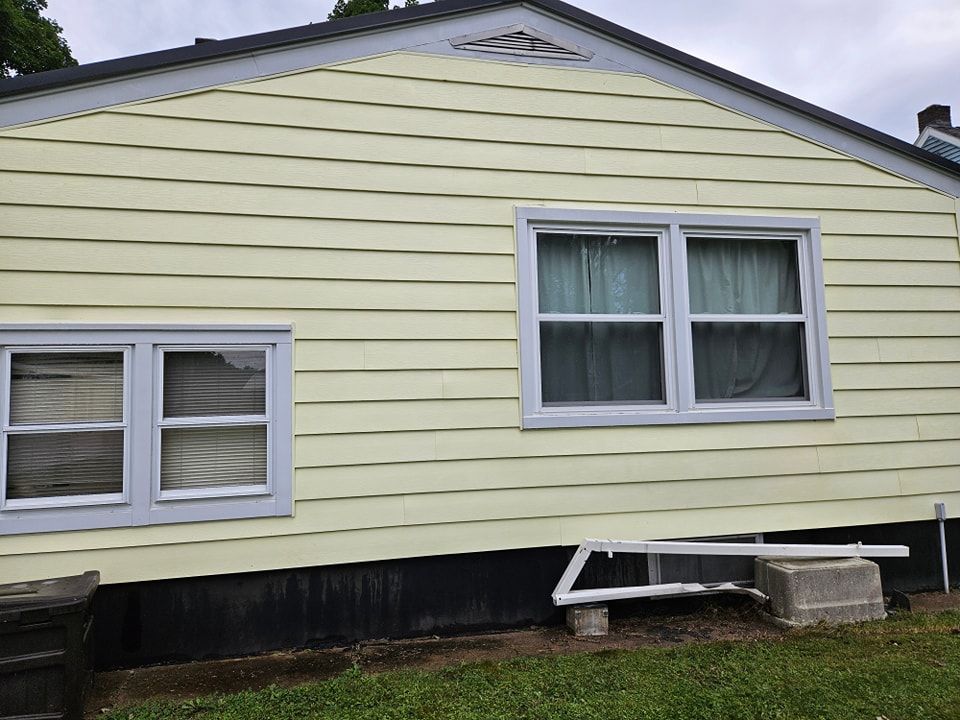 The exterior wall of a house with pale yellow siding, two windows, and a black foundation, with debris on the lawn.