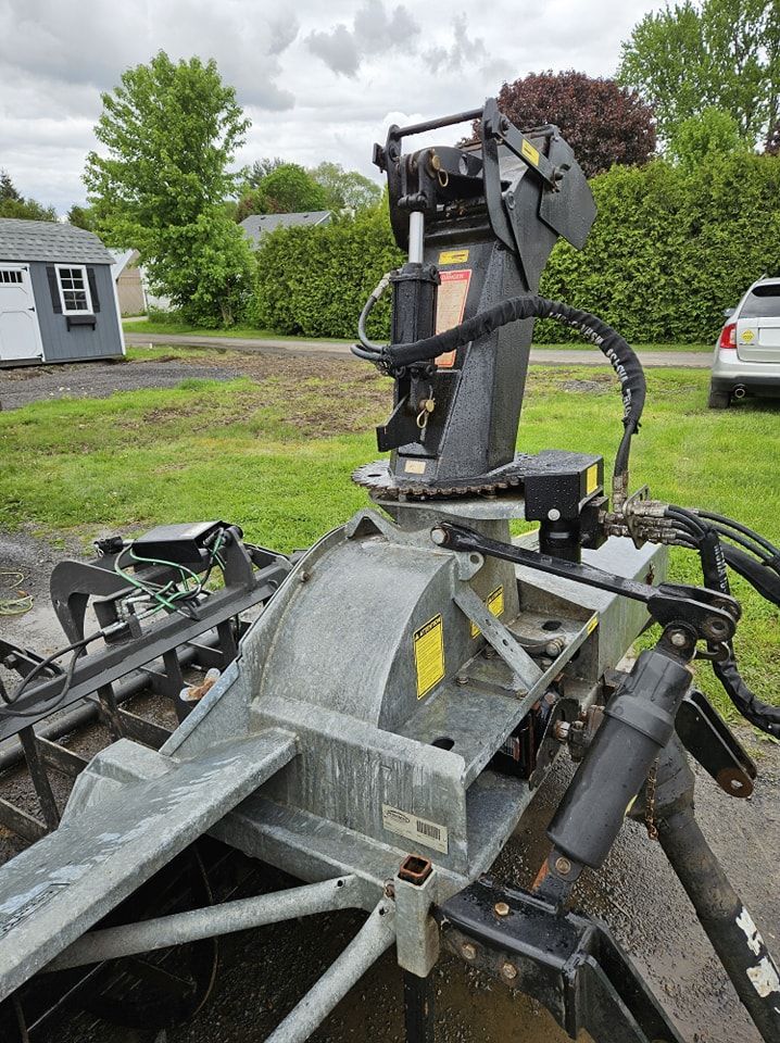 A metal snowblower attachment sits on a grassy lawn with a shed and car in the background.