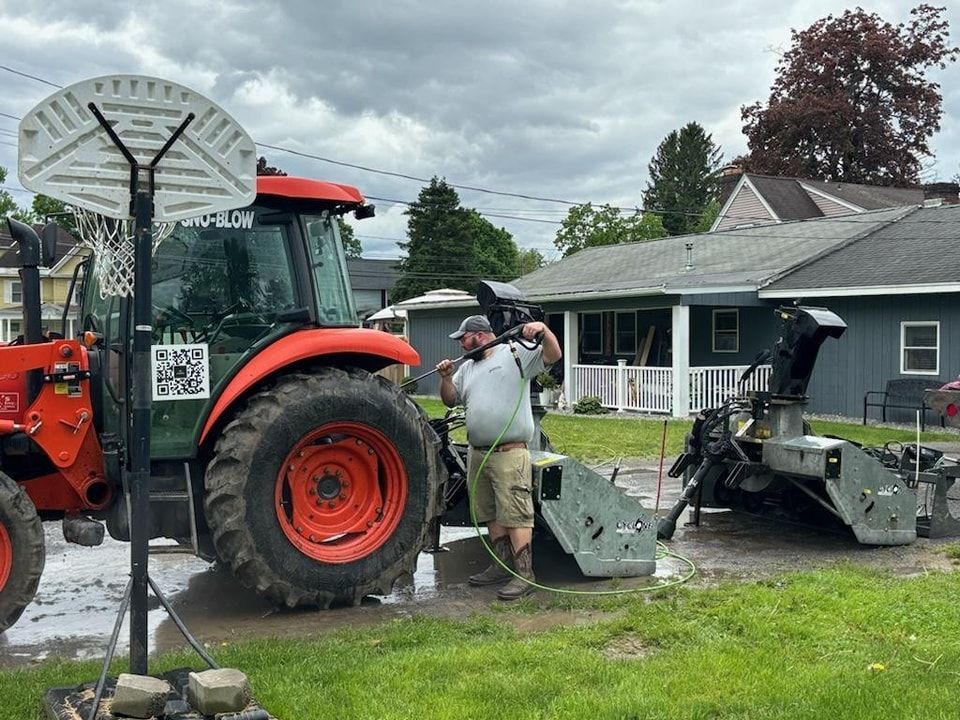 A person in a gray shirt and khaki shorts pressure washes a red tractor and snow blower attachment in a residential yard.