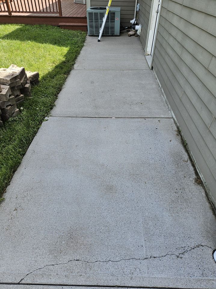 A gray concrete sidewalk runs alongside a house exterior with a small stack of stones on the lawn and a visible crack.