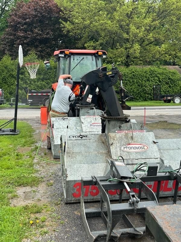 A person stands by an orange tractor and a Pronovost snow blower attachment on a gravel driveway near a basketball hoop.