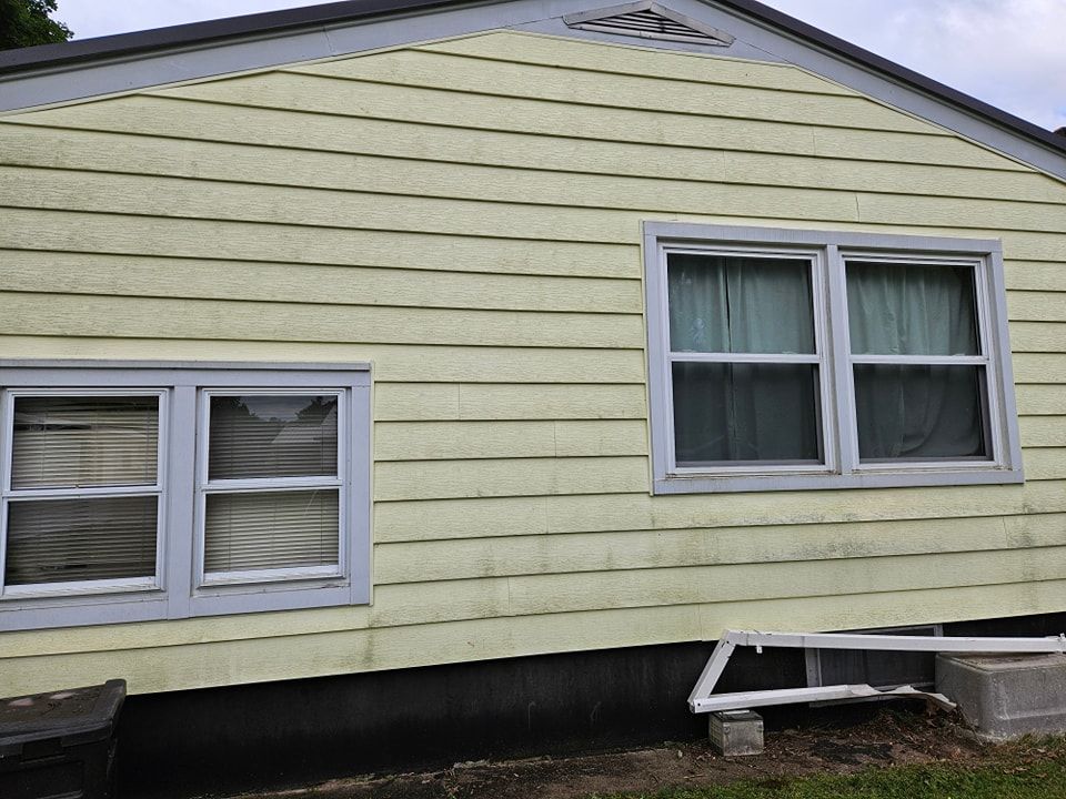 The side of a yellow-sided house with two grey-framed windows and a partially detached white gutter near the foundation.