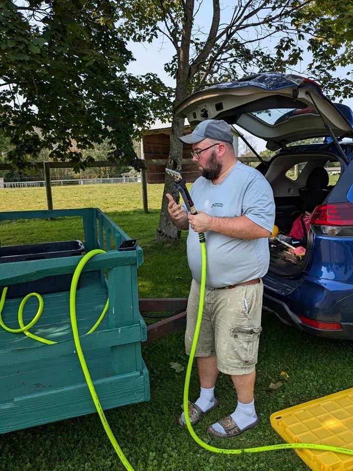 A person holding a lime-green hose next to a green utility trailer and a parked car outdoors on a grassy lawn.