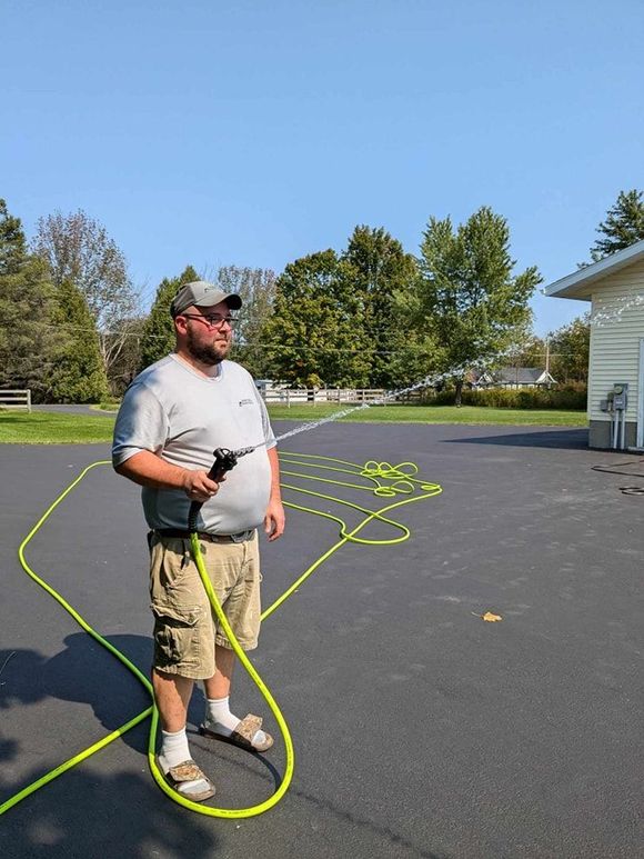 A person in a gray shirt, khaki shorts, and sandals stands on a paved driveway, spraying water from a neon green hose.