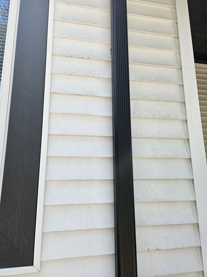 A black vertical downspout attached to white horizontal vinyl siding next to a dark window frame.