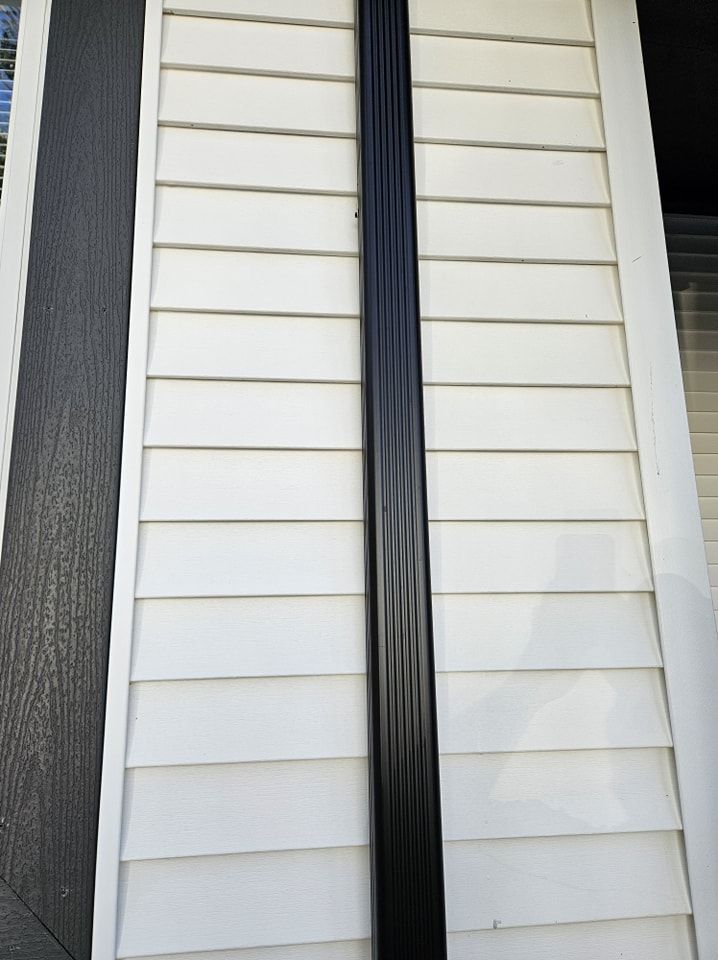 A black vertical downspout attached to a wall with horizontal white vinyl siding.