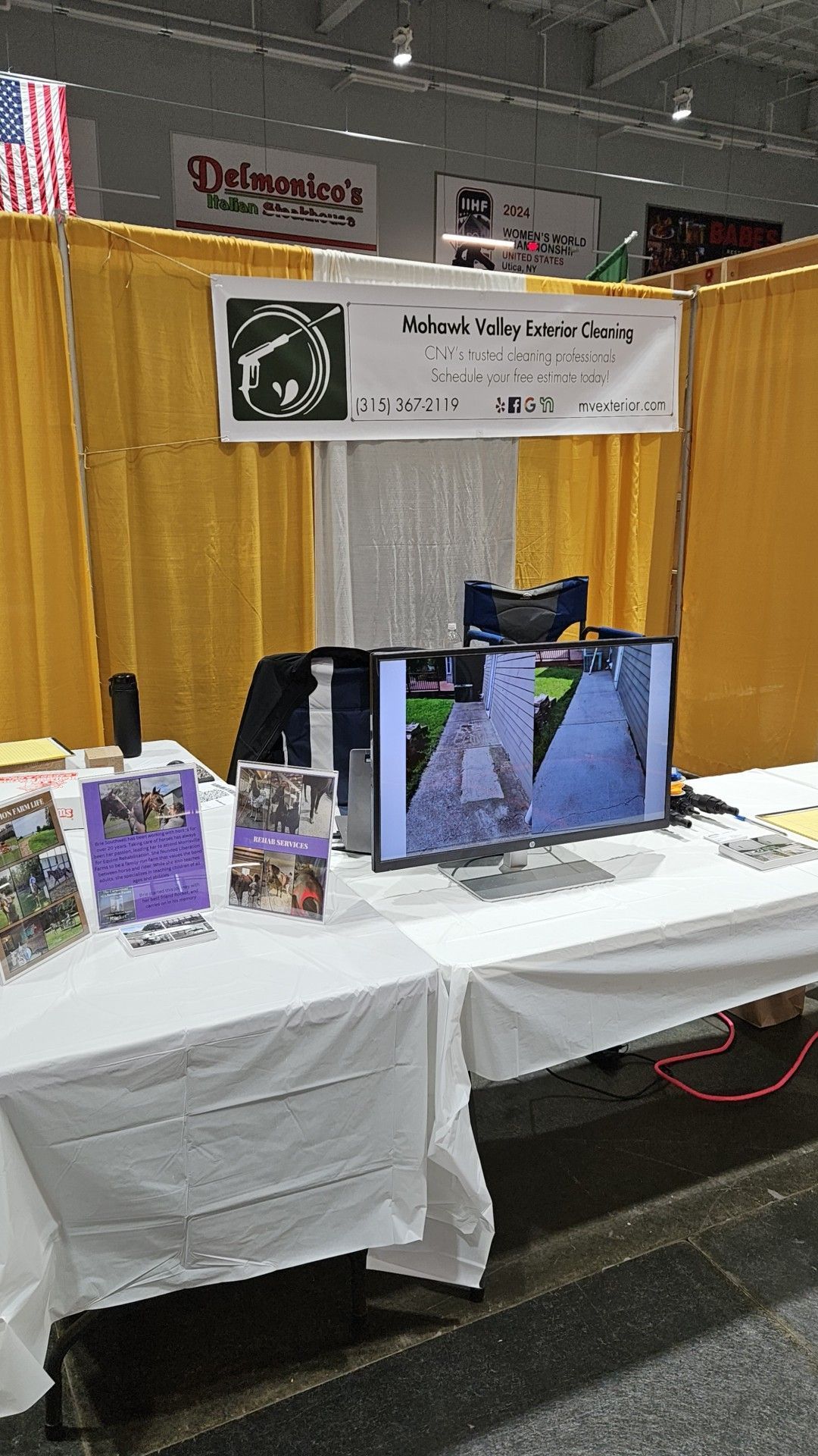 A trade show booth with yellow drapes, a white-covered table, brochures, and a monitor displaying video footage.