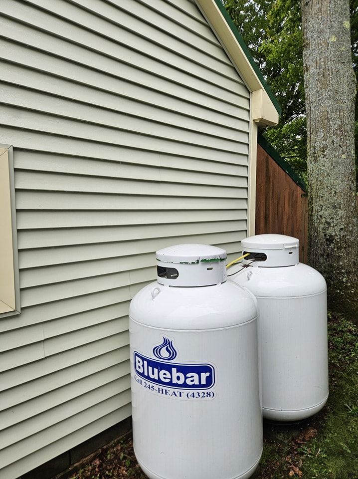 Two white propane tanks with Bluebar branding positioned against the beige vinyl siding of a house exterior.