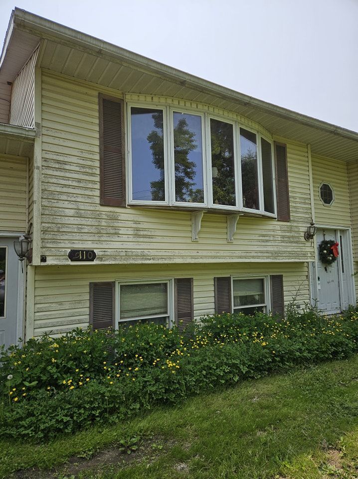 A yellow two-story house with a bay window, brown shutters, and overgrown landscaping in front.
