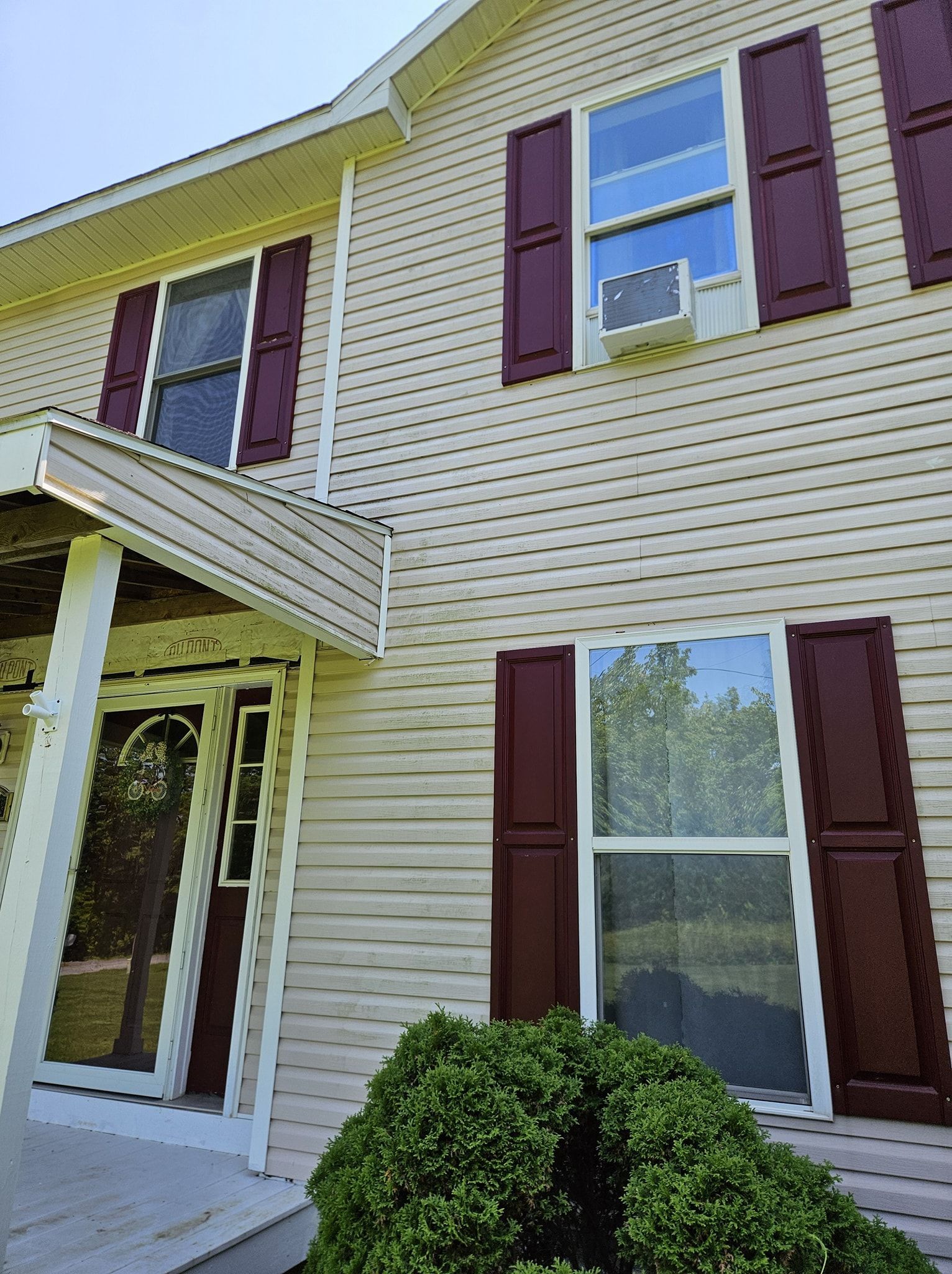 A two-story house with cream-colored siding, maroon shutters, a small front porch, and a window air conditioning unit.
